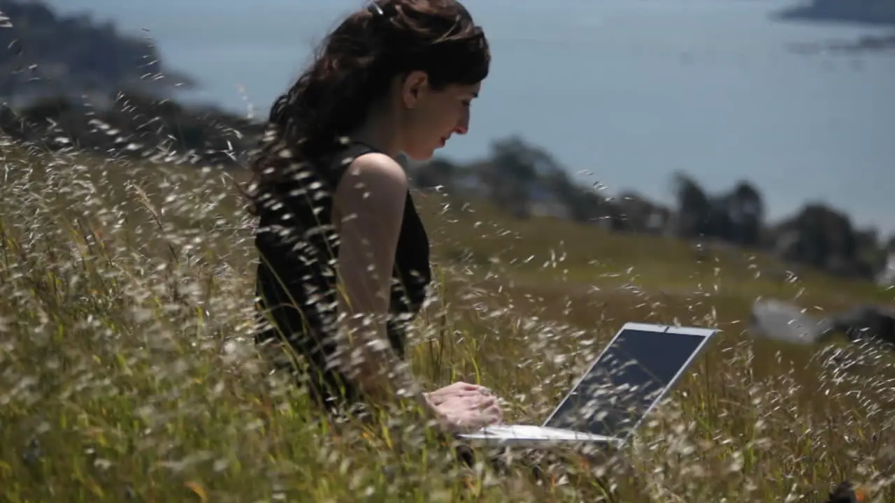 A woman using a laptop sits in a field by the shore