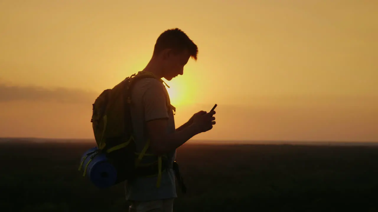 A Young Tourist Man With A Backpack Uses A Mobile Phone On Top Of A Mountain In The Rays Of The Sett