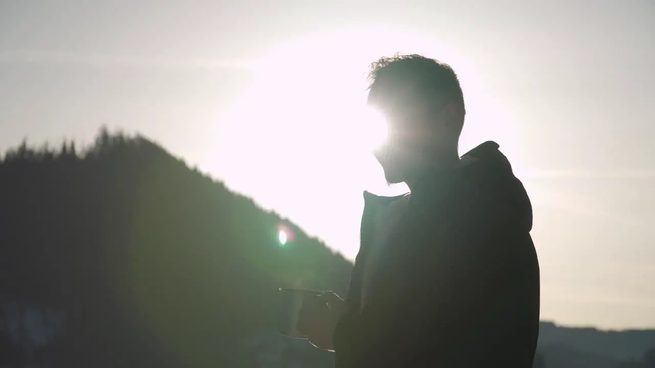 Happy smiling young man with coat drinks hot coffee or beverage with sun on horizon