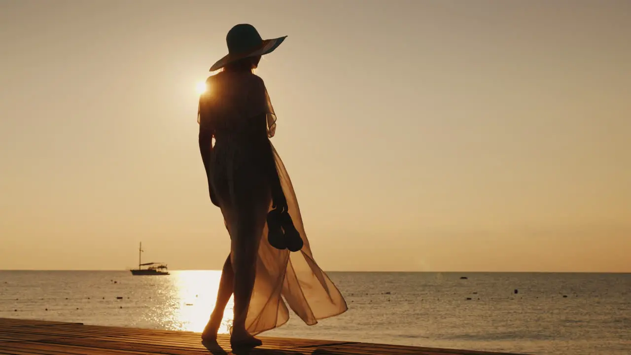 A Stylish Girl In Fluttering Clothes And A Broad-Brimmed Hat Meets The Sunrise On A Sea Pier