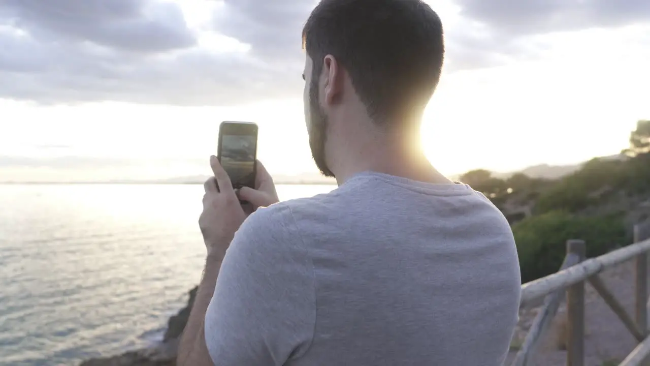 A man takes pictures of the sunset with the cell phone on the coast the sea and the golden sky are seen while he remains behind a fence inside a rural road