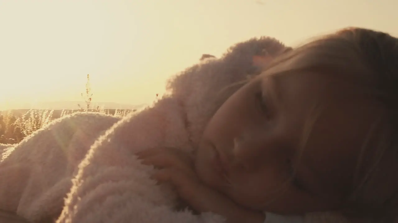 Pan around little girl snuggled in warm fuzzy jacket on picnic table as setting sun glows behind her