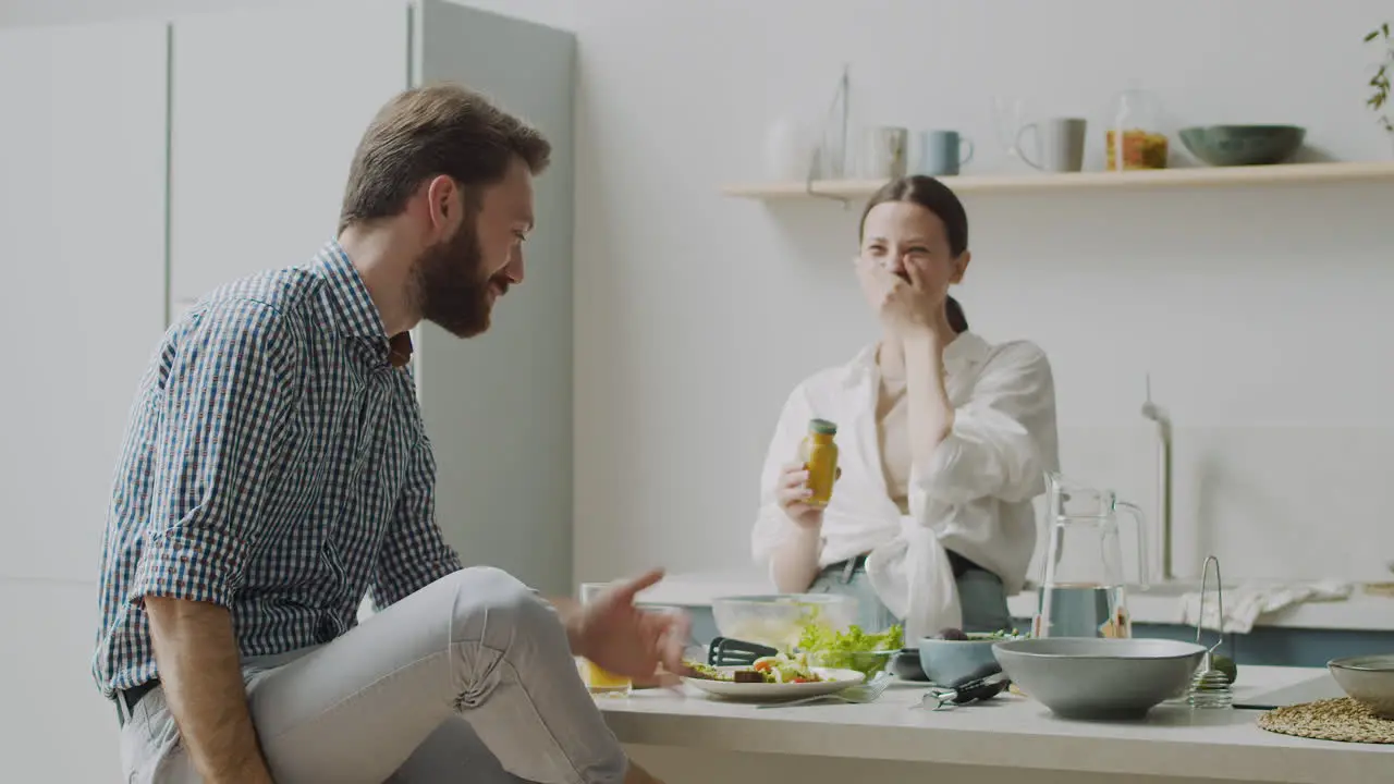 Happy Woman Standing Seasoning Salad While Man Sitting On Stool Telling Her Something Funny And Make Her Laugh