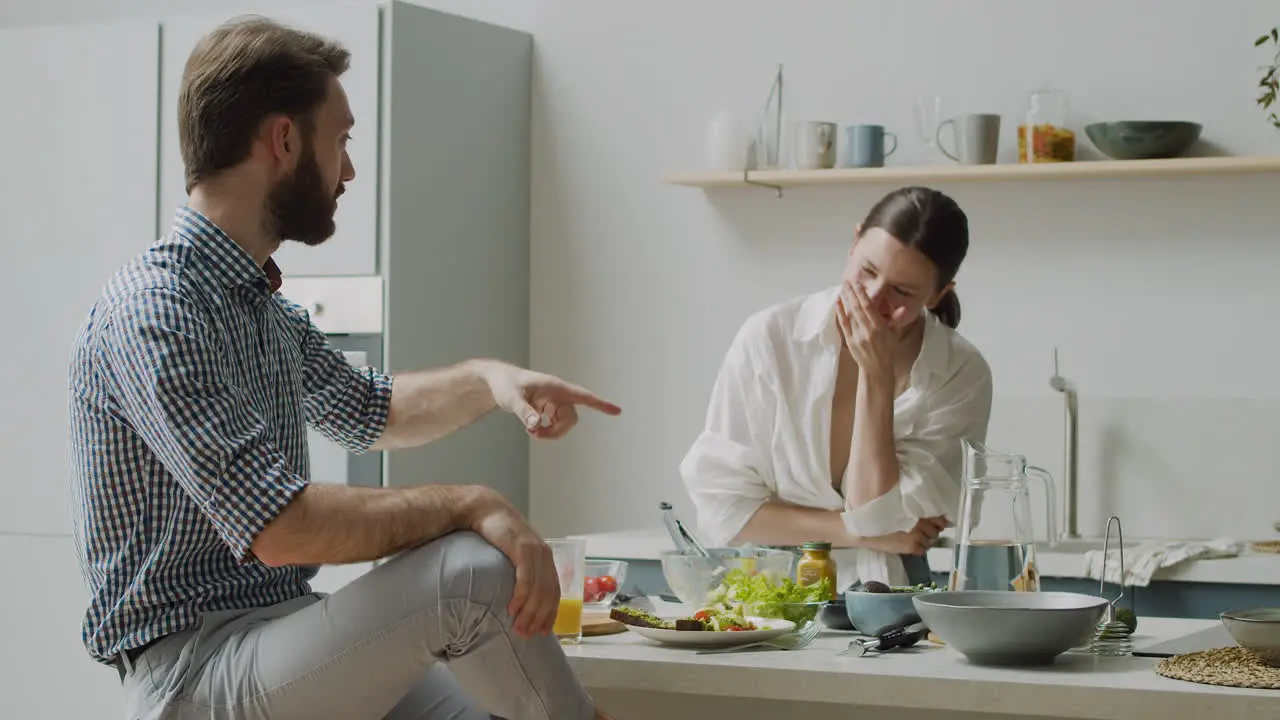 Beautiful Woman Mixing Salad In A Bowl While Her Boyfriend Sitting On A Stool Telling Her Something Funny And Make Her Laugh