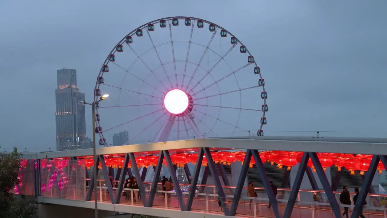 Nighttime view of a pedestrian bridge decorated with Chinese red lanterns as people walk through it while in the background there is a Ferris wheel in Hong Kong