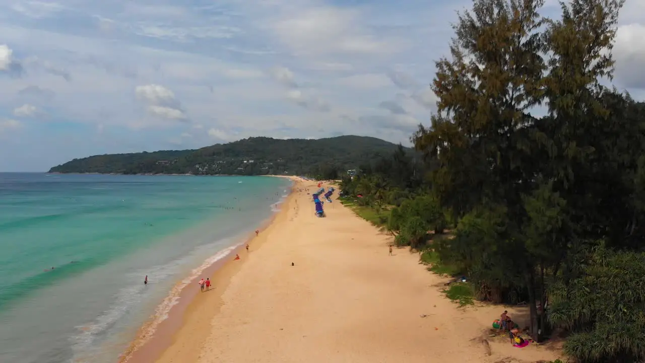 Pedestal drone shot of the length of Karon beachfront showing the scenic beauty of a sandy beach with crystal-clear waters against a backdrop of blue skies and lush green trees in Phuket Thailand