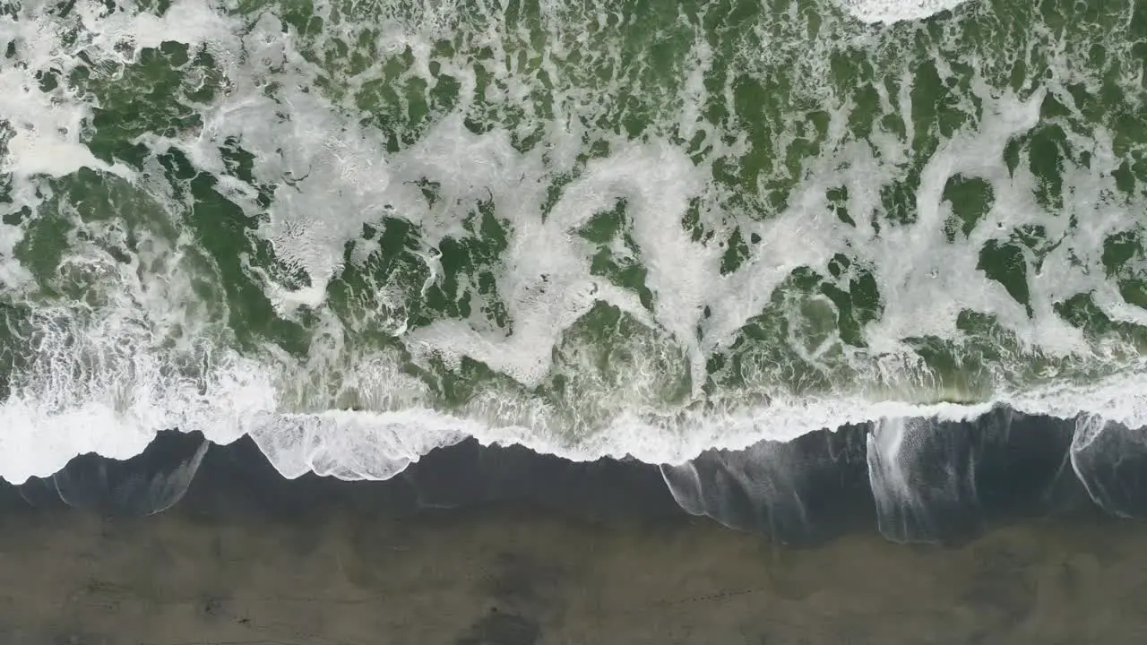 Aerial shot panning right over dark beach with green water and waves in Northern California