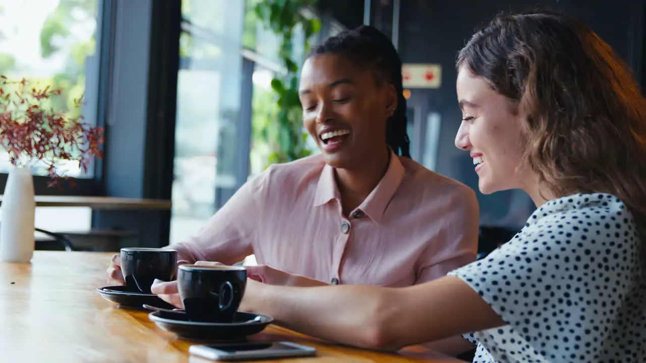 Two Young Female Friends Meeting In Coffee Shop And Looking At Mobile Phone