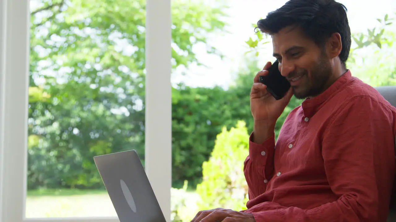 Man Working From Home Sitting On Floor In Lounge Using Laptop And Mobile Phone