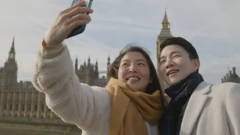 Young Asian Couple On Holiday Posing For Selfie In Front Of Houses Of Parliament In London UK