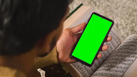 Close Up Of Muslim Man At Home Sitting On Floor And Reciting From The Quran Holding Green Screen Mobile Phone