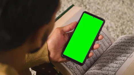 Close Up Of Muslim Man At Home Sitting On Floor And Reciting From The Quran Holding Green Screen Mobile Phone 1