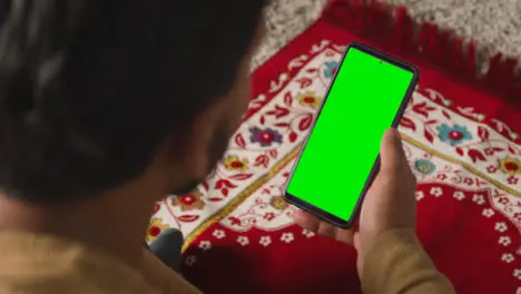 Close Up Of Muslim Man At Home Sitting On Floor On Prayer Mat Holding Green Screen Mobile Phone