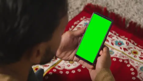 Close Up Of Muslim Man At Home Sitting On Floor On Prayer Mat Holding Green Screen Mobile Phone 1