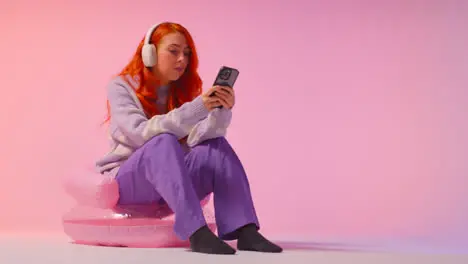 Studio Shot Of Young Gen Z Woman Wearing Headphones Streaming Music To Mobile Phone 3