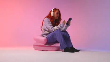 Studio Shot Of Young Gen Z Woman Wearing Headphones Streaming Music To Mobile Phone