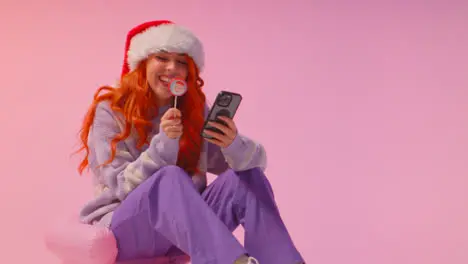 Studio Shot Of Young Gen Z Woman Wearing Christmas Santa Hat Eating Candy Lollipop Looking At Mobile Phone 3