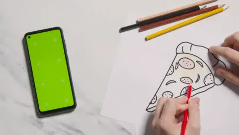 Overhead Shot Of Person Colouring In Picture Of Pizza With Green Screen Phone On Marble Background