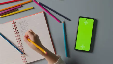 Overhead Shot Of Child With Green Screen Mobile Phone Doing School Maths Homework In Book 1