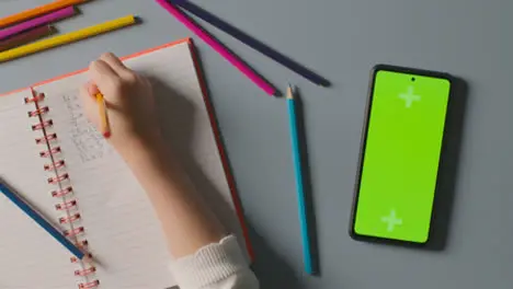 Overhead Shot Of Child With Green Screen Mobile Phone Doing School Maths Homework In Book