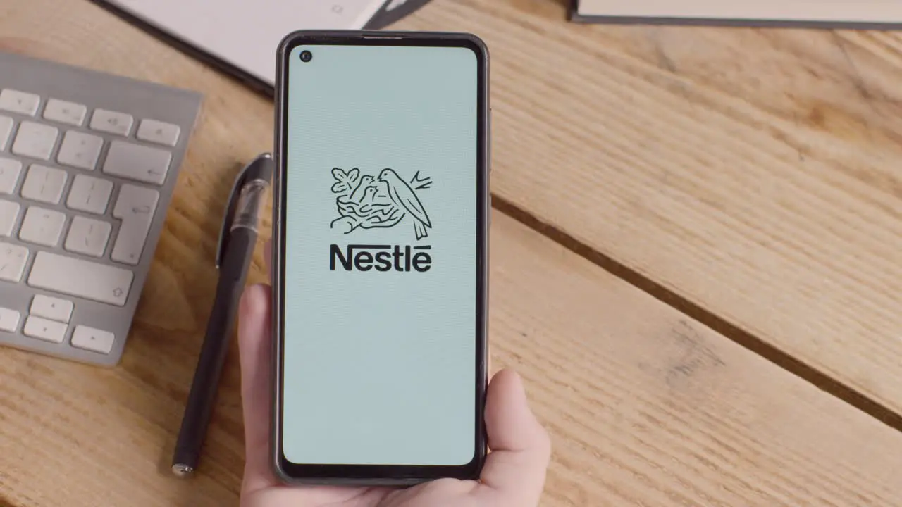 Person Holding Mobile Phone On Desk With Screen Showing Brand Logos For McDonalds Nestle Coca Cola Starbucks And Pepsico