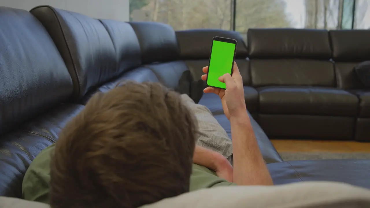 Man wearing pyjamas lying on sofa at home looking at green screen mobile phone shot in slow motion