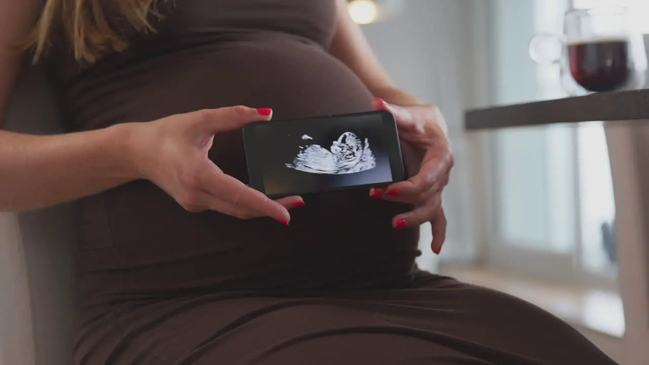 Close Up Of Pregnant Woman At Home Holding Mobile Phone With Ultrasound Scan Of Baby