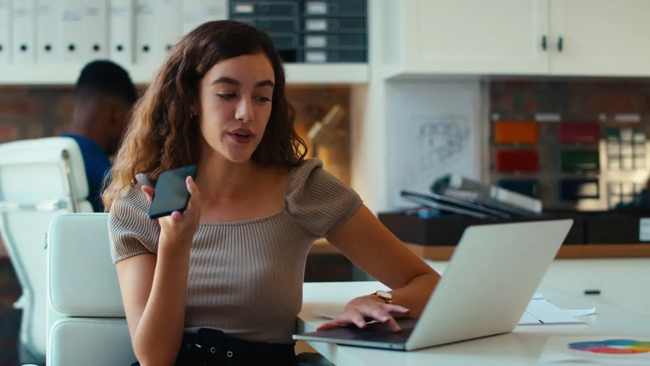 Young Smiling Businesswoman Working At Desk In Office Talking Into Mic Of Mobile Phone