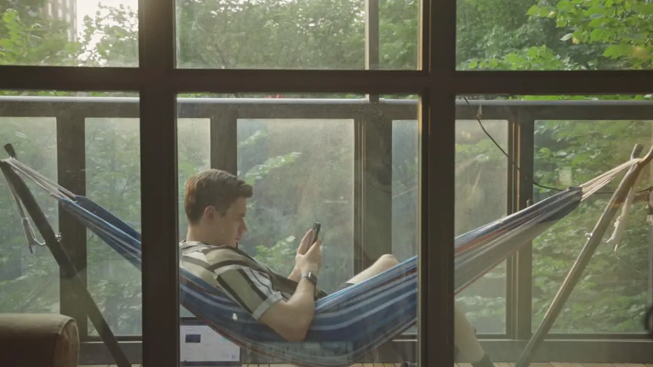 A Man Ridding In A Hammock While Using Cellphone Outside The House