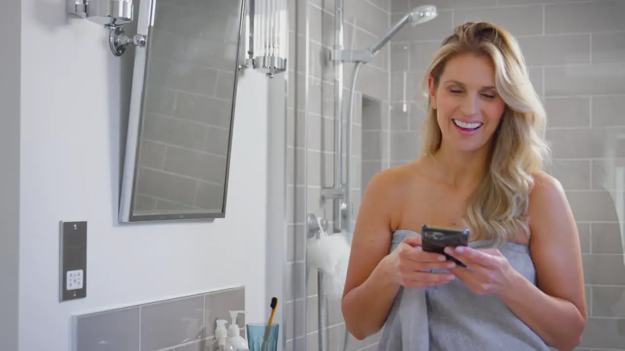 Mature Woman Getting Ready In Bathroom At Home Looking At Mobile Phone And Laughing