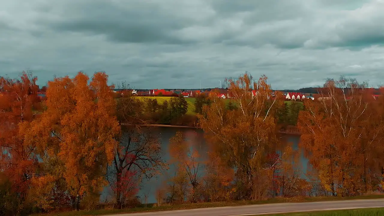 a panorama cloudy sky red roofs of a small town meadow and beautiful trees with autumn colors along the lake