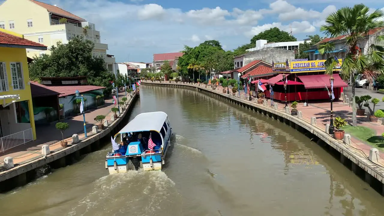 Narrow looking Malacca River with houses while boat is cruising the river and a cloudy blue sky in the background