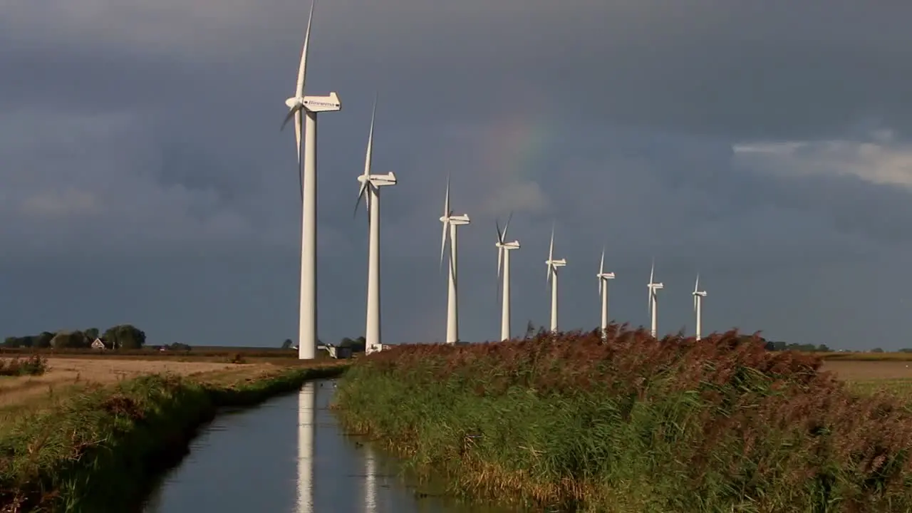 A line of Wind Turbines turning with a drainage ditch in the foreground and a rainbow and stormy sky