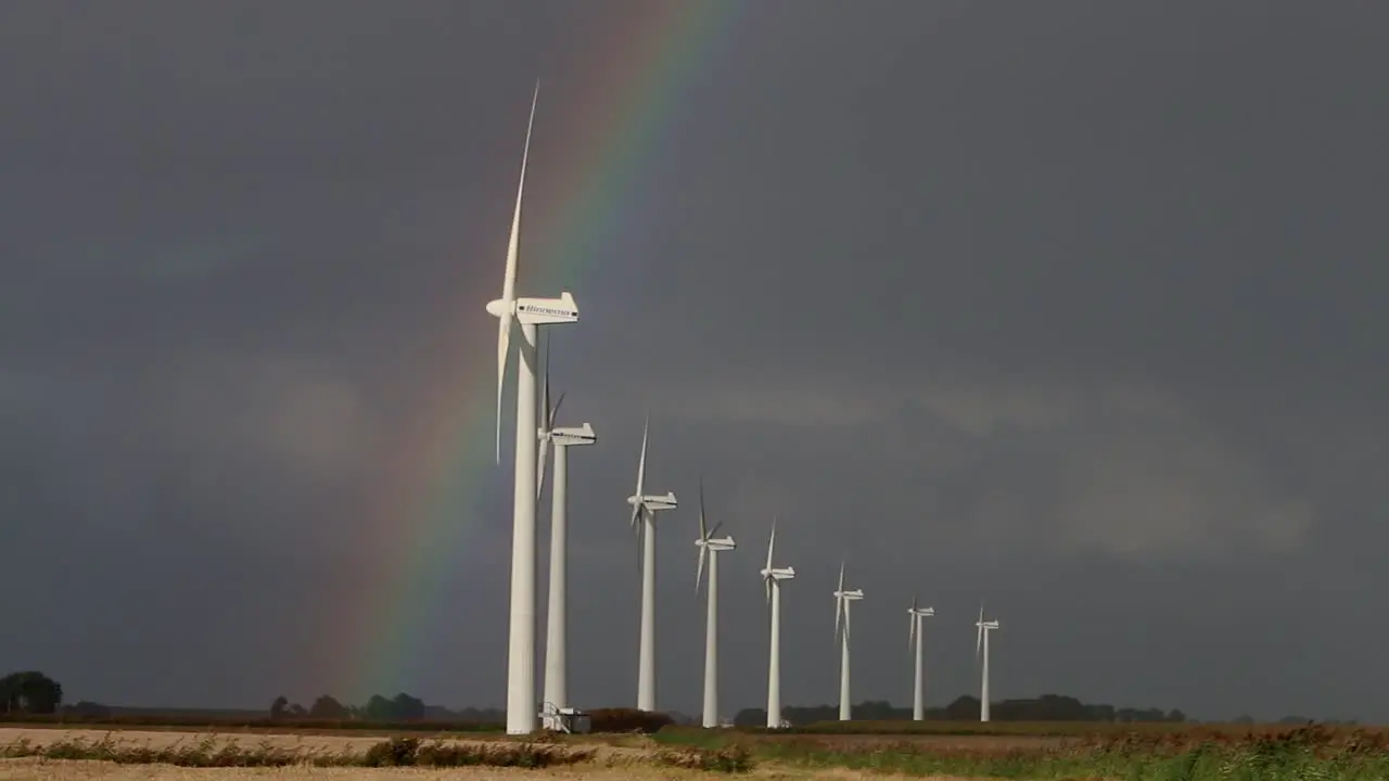 A line of Wind Turbines turning on a windy day with a stormy sky and rainbow in the background