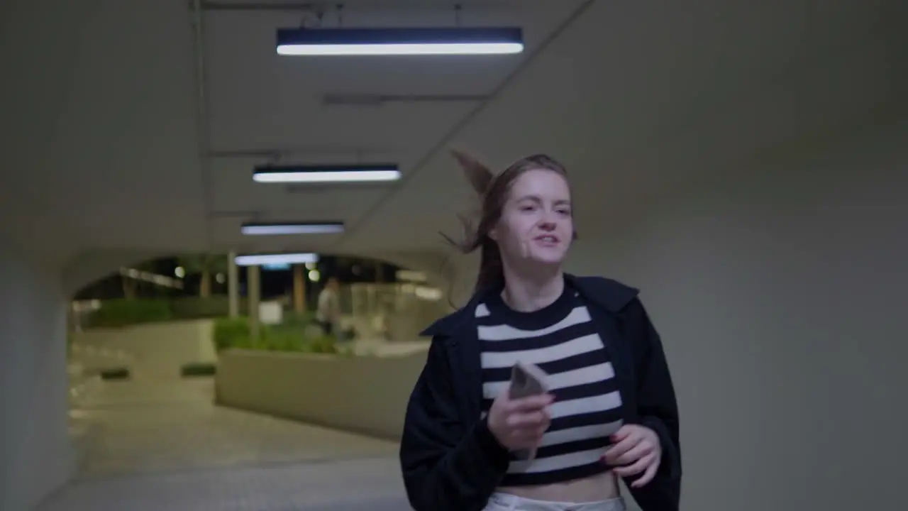 A person with wavy hair runs through a luminous tunnel in a skateboard park during the night passing in front of the camera