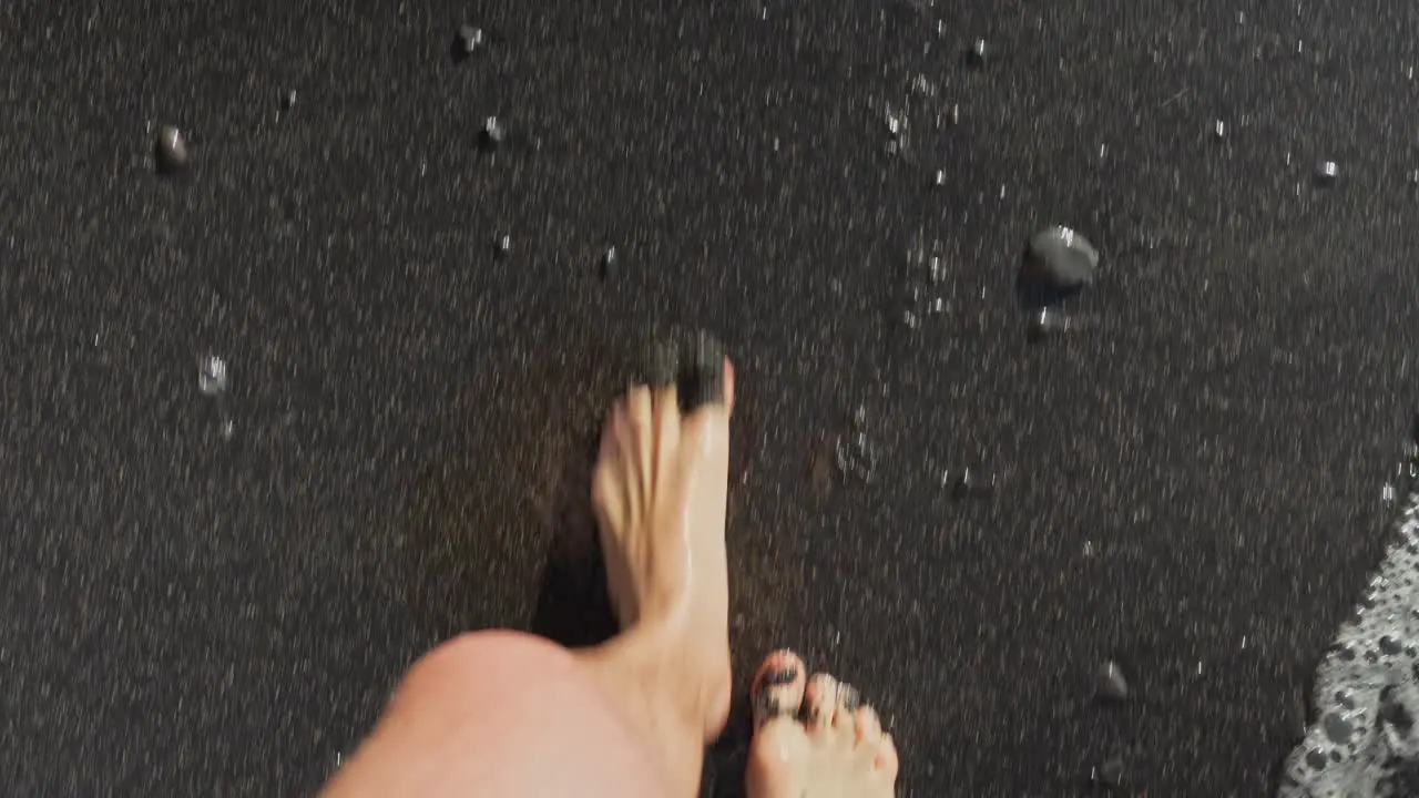 Top down POV of person walking along black pebble sand beach as ocean waves recede