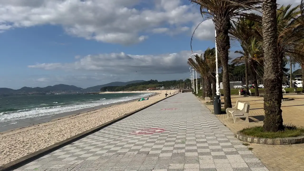 Pov walk along promenade of Vigo CIty with palm trees sandy beach and strong ocean waves