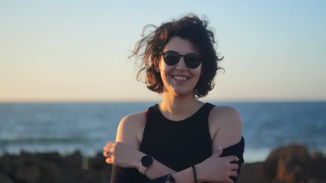 Young woman wearing black shades smiling at the camera ocean behind her at sunset