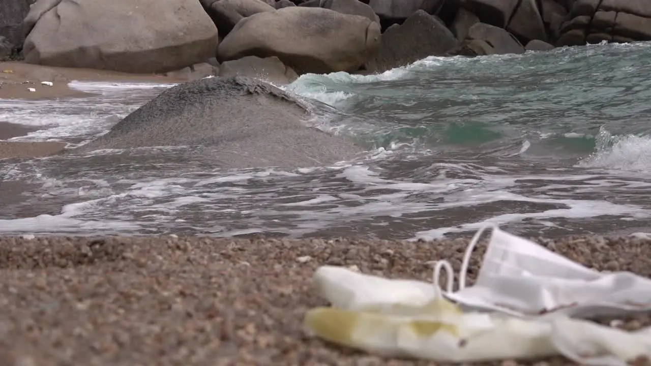 Polluted mask and glove on a beach with rushing wave nearby static shot focusing background