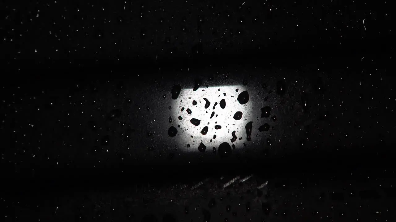 A street light at night is seen out of focus through rain drops dripping down a misted window that is behind a Venetian blind