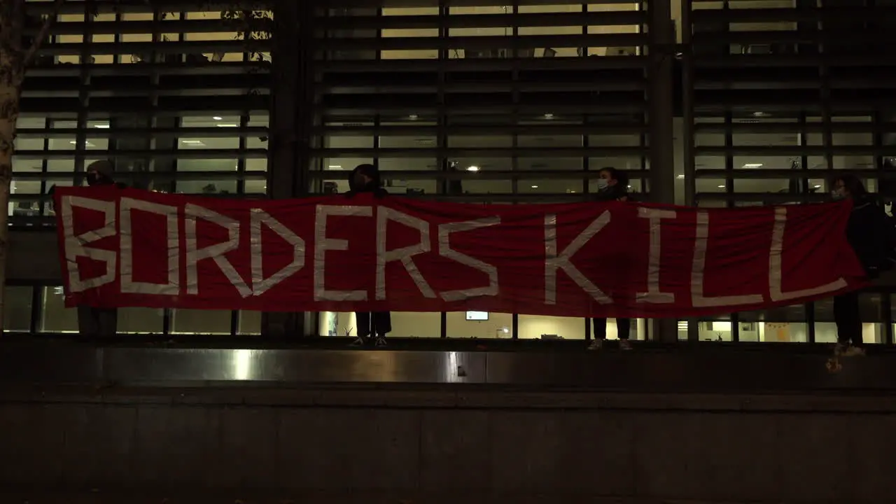 Protestors hold up a large red banner that says Borders Kill” outside the UK Home Office at night on a protest for refugee rights