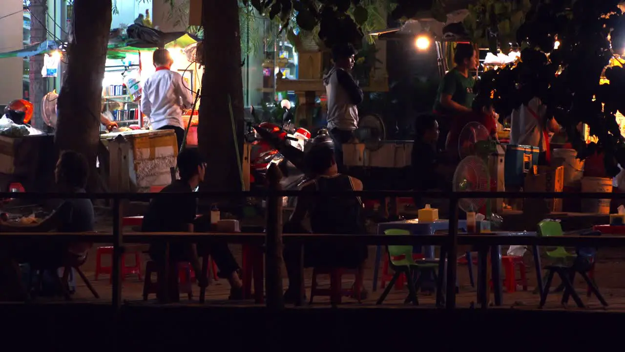 Wide Exterior Shot of People Sitting on Small Plastic Chairs and Talking in a Small Out Door Restaurant wile Others Are Busy In The Background in the Night Time