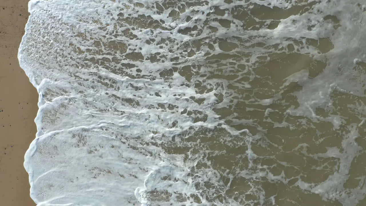 Aerial shot of big powerful high white foam waves crashing on a golden sand beach