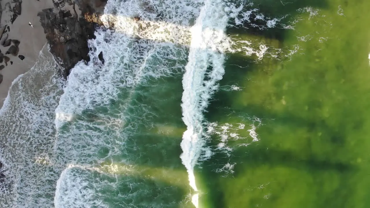 A lime green ocean mixed with hints of dark blue pushes small waves into the dark rocks sat between the pale coarse sand whilst shadows cast from apartment buildings block the sunsets afternoon glow