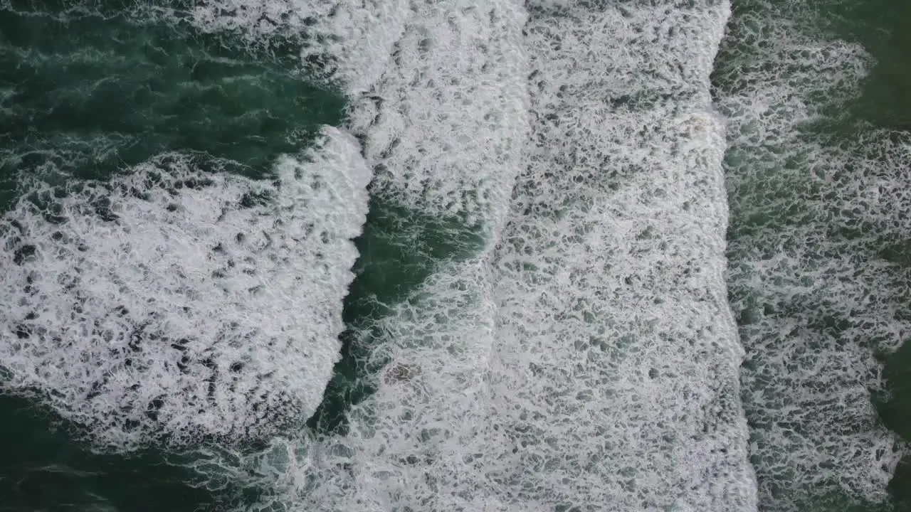 A top down cinematic drone shot of green Cornish waves ideal for surfing slowly rolling and breaking on the shore