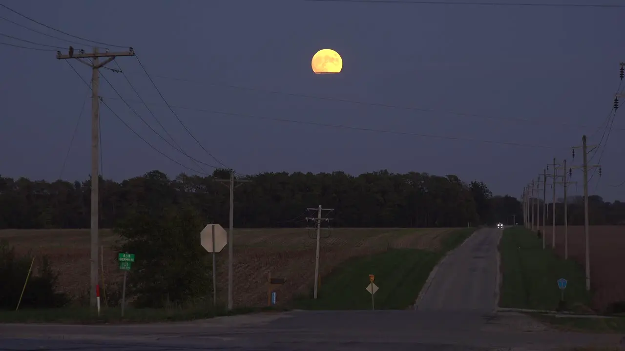 A Distant Car Drives Down A Lonely Country Road At Night With A Full Moon Rising