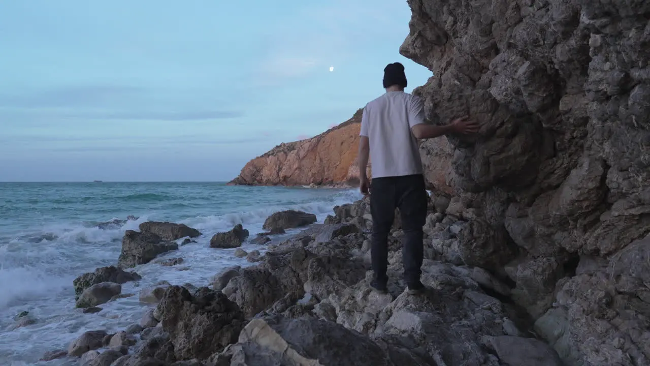 man walks on rocks near cliffs as sea washes onshore moon shines above in blue sky he takes a moment to reflect