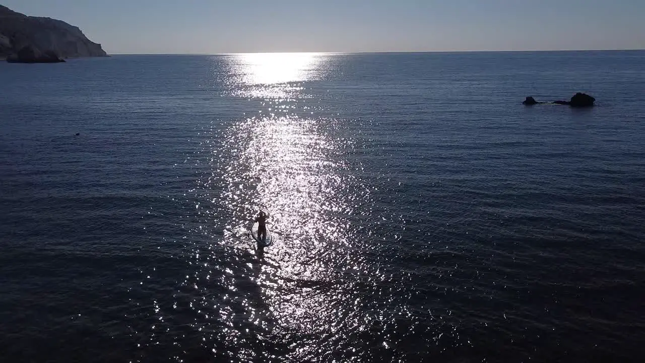 Beautiful sunrise aerial view of a person paddleboarding out to sea as the sun's reflection creates a sparkling path towards the open water