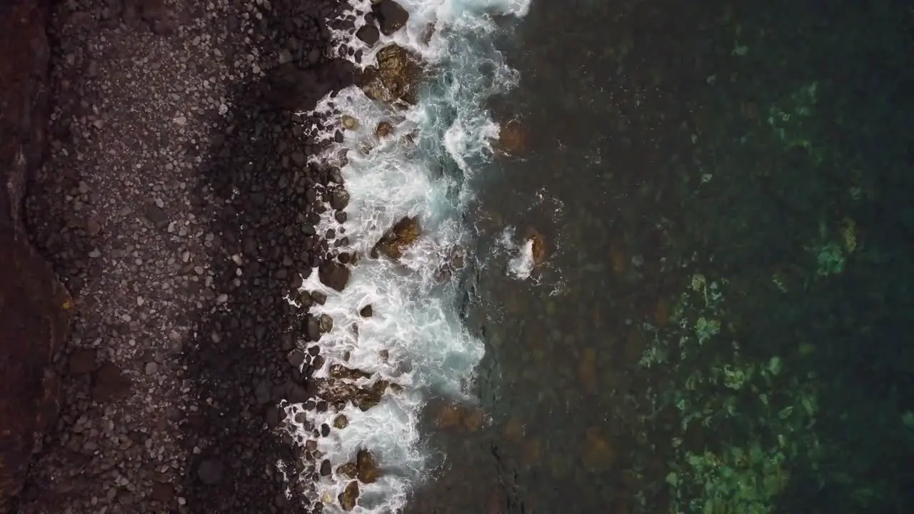 Top down aerial relaxing view of volcanic rocky beach coast while waves breaks on shore