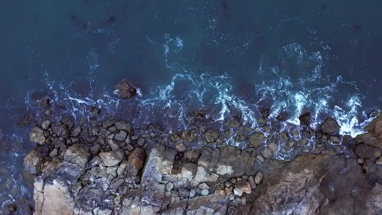 Waves gently breaking against a rocky shoreline straight down aerial view